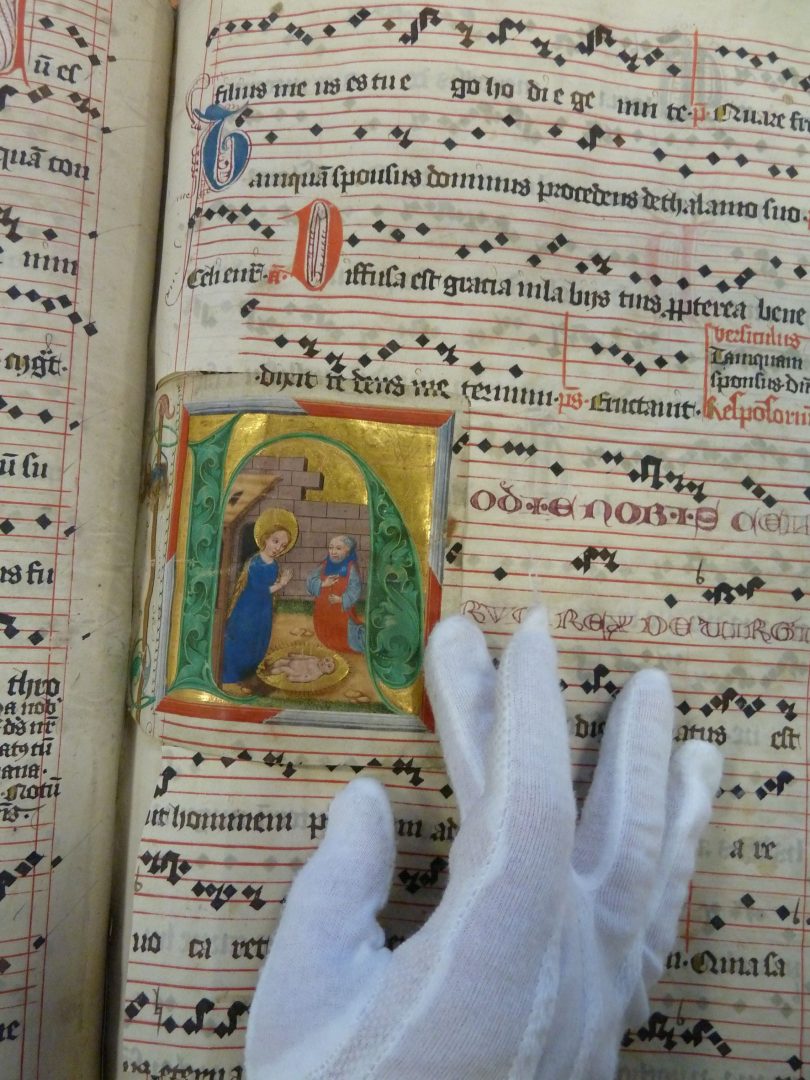 Karta pergaminowa z ozdobnymi literami; na niej kolorowy obrazek z postacią świętej trzymany przez rękę w białej rękawiczce