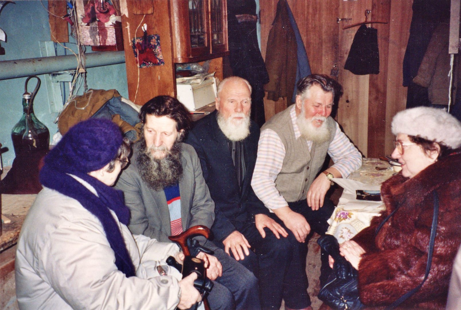 Trzech starszych mężczyzn z brodami siedzi w pomieszczeniu mieszkalnym i rozmawia z kobietą w ciepłym płaszczu