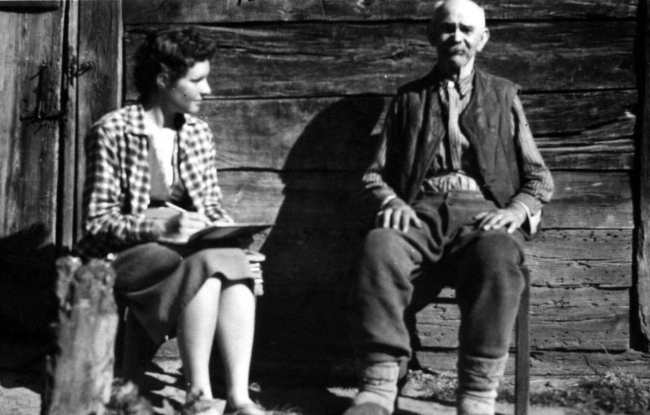 Czarno-biała fotografia; kobieta z notatnikiem i starszy mężczyzna siedzą na tle drewnianej ściany