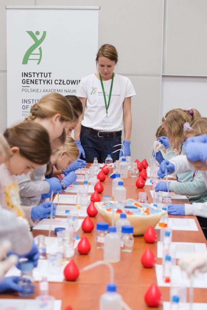 Kobieta stoi u szczytu długiego stołu przy którym siedzą dzieci zajęte doświadczeniami chemicznymi