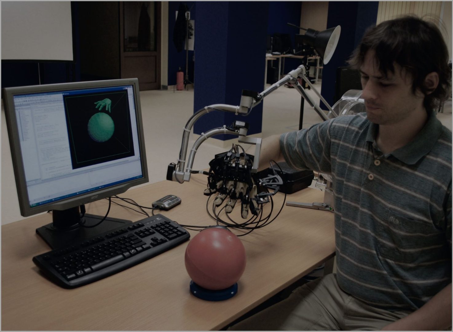 Mężczyzna ma na prawej ręce nałożoną elektroniczną rękawicę, którą zbliża do czerwonej piłki; na monitorze komputera odwzorowanie tego ruchu