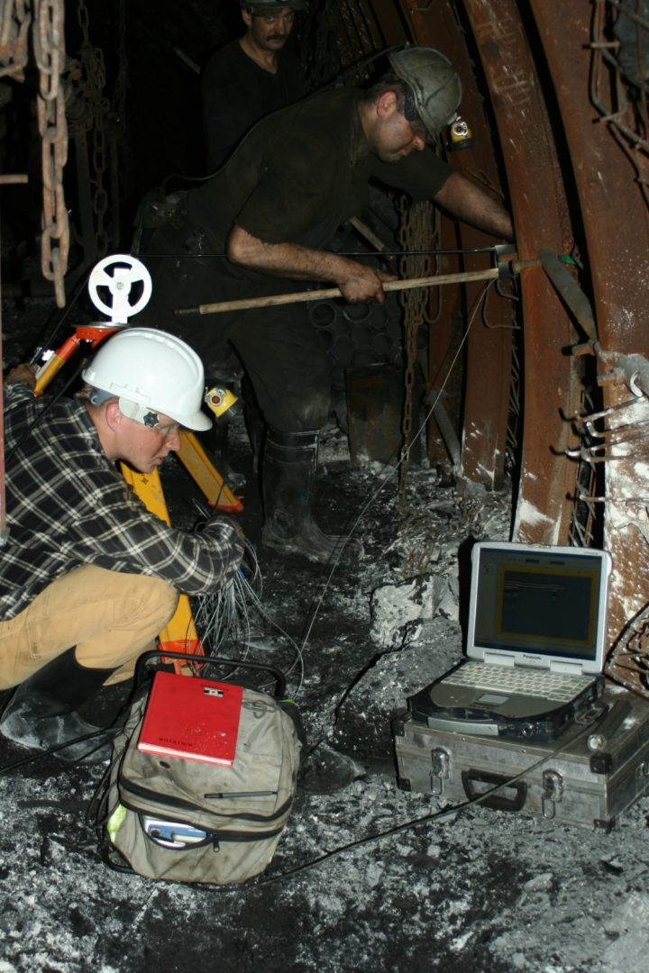 Kopalnia; jeden mężczyzna dociska do ściany długi drąg, drugi spogląda w leżący na ziemi laptop