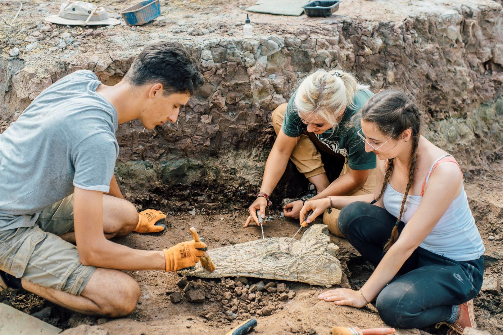 Troje młodych ludzi pędzelkami oczyszcza wykopalisko na stanowisku archeologicznym