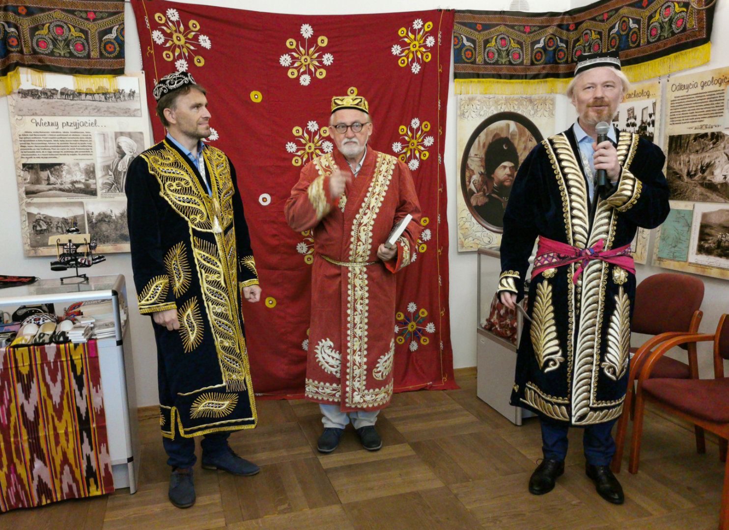 Uczestnicy wernisażu (trzech mężczyzn) opowiadają o wystawie historyczno-przyrodniczej o Pamirze w regionalnych strojach i nakryciach głowy na tle udekorowanej tkaninami, obrazami i zdjęciami sali