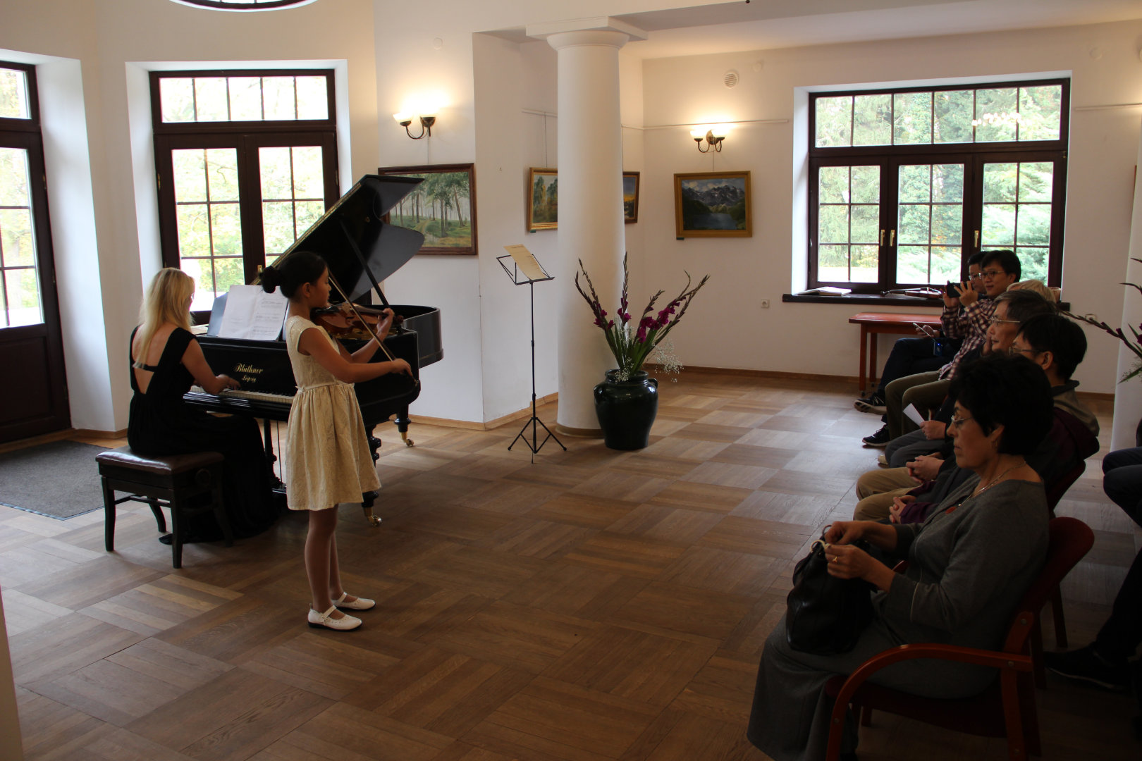 W willi Janówek dziewczynka gra na skrzypcach, której akompaniuje na fortepianie nauczycielka podczas koncertu uczniów szkoły muzycznej, obok widownia